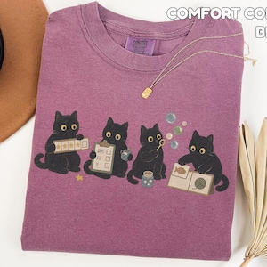 May include: A berry-colored Comfort Colors t-shirt featuring a graphic of four black cats. One cat holds a five-star rating card, another a clipboard, the third blows bubbles, and the fourth examines a fish and yarn card. The text reads Comfort Colors Berry.