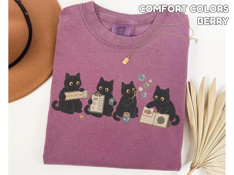 May include: A berry-colored Comfort Colors t-shirt featuring a graphic of four black cats. One cat holds a five-star rating card, another a clipboard, the third blows bubbles, and the fourth examines a fish and yarn card. The text reads Comfort Colors Berry.