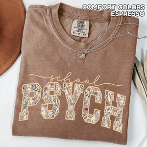 May include: Espresso-colored t-shirt with the words "school PSYCH" in a floral design. The shirt has a crew neck and short sleeves. A silver necklace with a pendant is draped across the shirt.