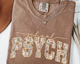 May include: Espresso-colored t-shirt with the words "school PSYCH" in a floral design. The shirt has a crew neck and short sleeves. A silver necklace with a pendant is draped across the shirt.