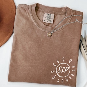 May include: A brown t-shirt with a crew neck and the text "SLP" inside a sun graphic. The shirt has a small tag that says "Comfort Colors". A silver necklace with a pendant is draped across the shirt.