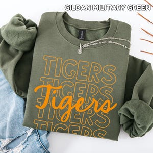 May include: A military green sweatshirt with the word "Tigers" in orange block letters and a cursive font. The sweatshirt is folded on top of a pair of blue jeans. A silver necklace with a pendant is on the sweatshirt.