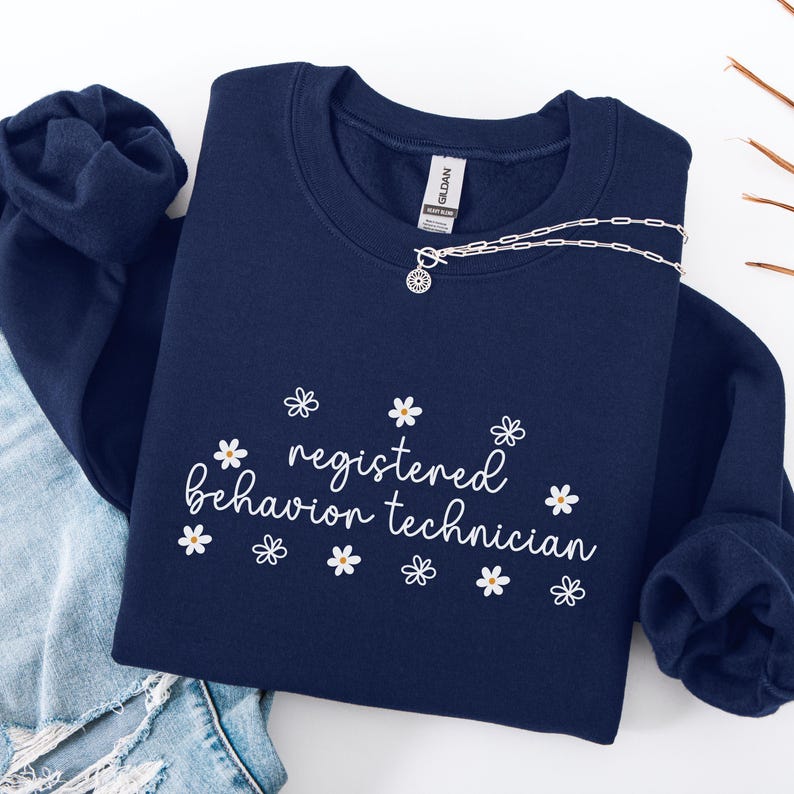 K&ouml;nnte beinhalten: Navyblauer Sweatshirt mit dem Text "registered behavior technician" in wei&szlig;er Schrift mit wei&szlig;en G&auml;nsebl&uuml;mchen-Akzenten.