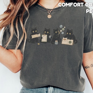 May include: A charcoal gray short-sleeve t-shirt featuring a graphic of four black cats engaged in various activities, including holding a checklist and blowing bubbles. The text COMFORT COLORS PEPPER is printed in the upper right corner.