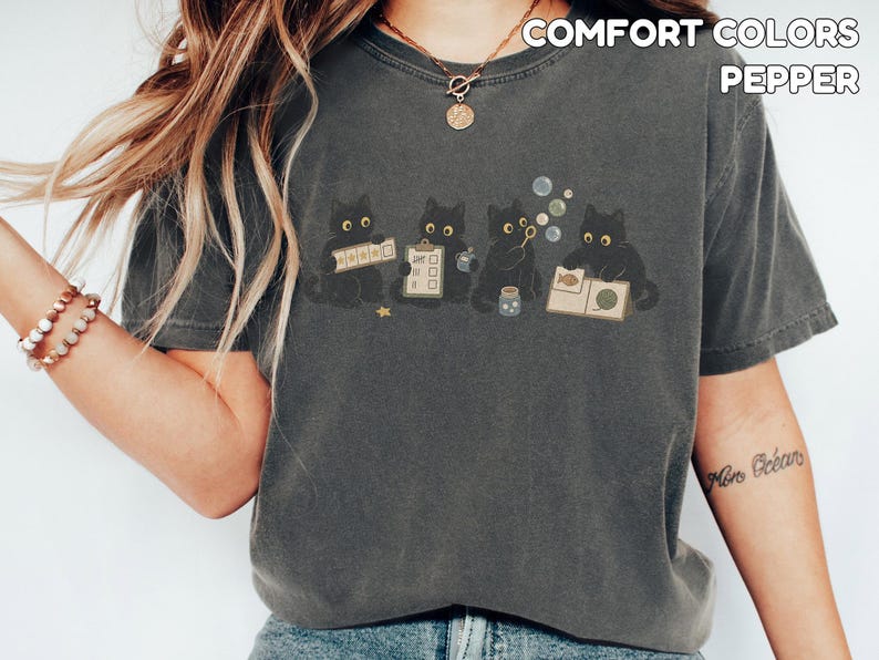 May include: A charcoal gray short-sleeve t-shirt featuring a graphic of four black cats engaged in various activities, including holding a checklist and blowing bubbles. The text COMFORT COLORS PEPPER is printed in the upper right corner.