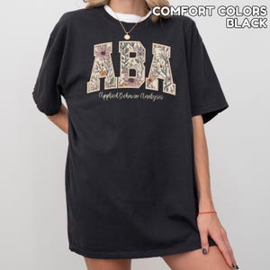 May include: Black t-shirt with the letters "ABA" in a floral design. The words "Applied Behavior Analysis" are printed below the letters. The shirt is a comfort colors style.