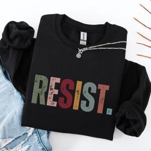 Pode incluir: Camisola de moletom preta com a palavra "RESIST" impressa em letras coloridas de estilo vintage.