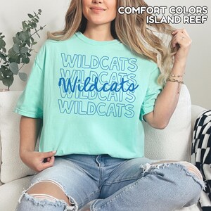 May include: A light green t-shirt with the word "Wildcats" printed in blue multiple times. The shirt is a solid color and has short sleeves. The text "Comfort Colors Island Reef" is printed at the top.