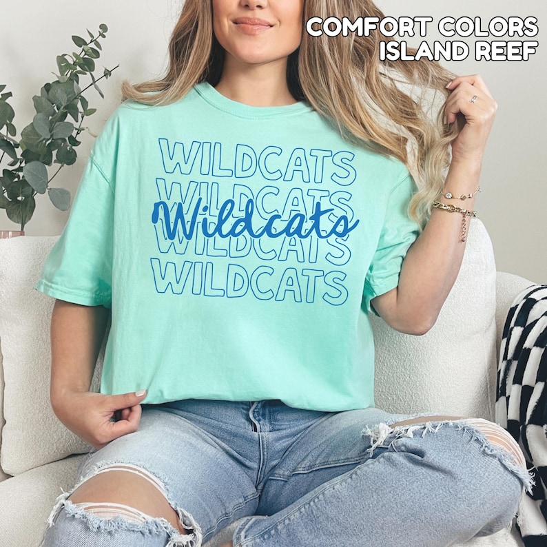 May include: A light green t-shirt with the word "Wildcats" printed in blue multiple times. The shirt is a solid color and has short sleeves. The text "Comfort Colors Island Reef" is printed at the top.
