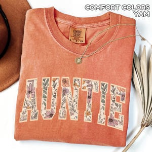 Puede incluir: Una camiseta de color coral con la palabra "AUNTIE" en letras grandes y florales. La camiseta tiene cuello redondo y una peque&ntilde;a etiqueta en el cuello. Un collar de oro con un colgante est&aacute; drapeado sobre la camiseta. El texto "COMFORT COLORS YAM" est&aacute; en la parte superior.