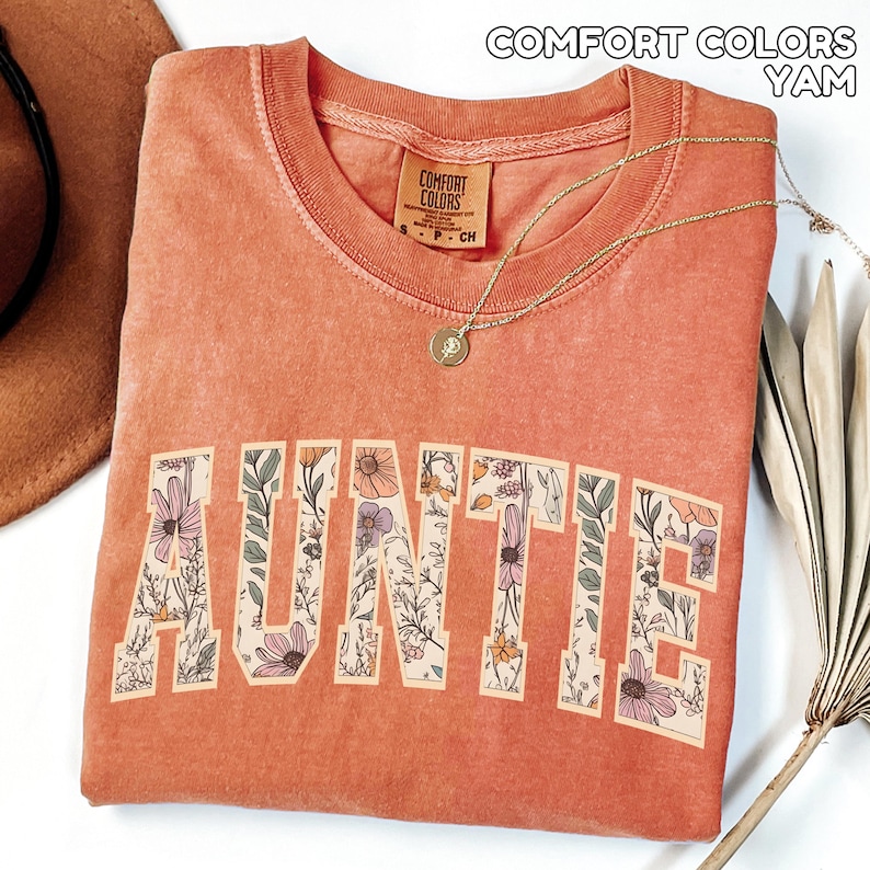 Puede incluir: Una camiseta de color coral con la palabra "AUNTIE" en letras grandes y florales. La camiseta tiene cuello redondo y una peque&ntilde;a etiqueta en el cuello. Un collar de oro con un colgante est&aacute; drapeado sobre la camiseta. El texto "COMFORT COLORS YAM" est&aacute; en la parte superior.
