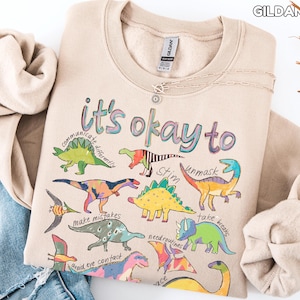 May include: A sand-colored sweatshirt with the text "it's okay to" and various dinosaur illustrations. The sweatshirt also features phrases like "communicate differently" and "unmask". A silver necklace is draped across the front.
