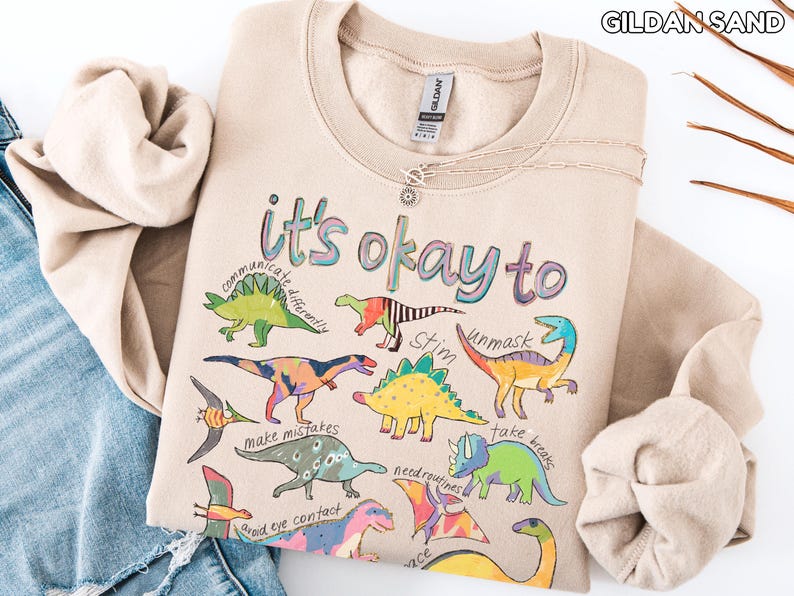 May include: A sand-colored sweatshirt with the text "it's okay to" and various dinosaur illustrations. The sweatshirt also features phrases like "communicate differently" and "unmask". A silver necklace is draped across the front.