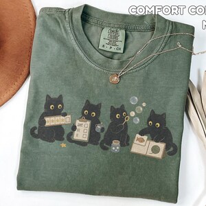 May include: A moss green t-shirt featuring a graphic of four black cats. One cat holds a five-star rating card, another a clipboard, one blows bubbles, and the fourth looks at a book with a fish and yarn illustration. The shirt has a Comfort Colors label.