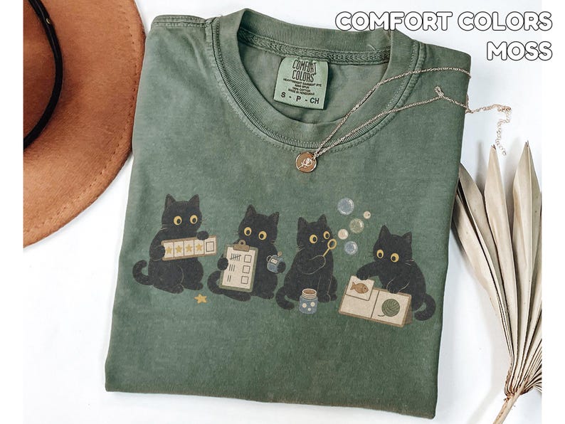 May include: A moss green t-shirt featuring a graphic of four black cats. One cat holds a five-star rating card, another a clipboard, one blows bubbles, and the fourth looks at a book with a fish and yarn illustration. The shirt has a Comfort Colors label.
