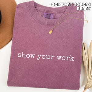K&ouml;nnte beinhalten: Beerenfarbenes Comfort Colors T-Shirt mit dem wei&szlig;en Schriftzug "show your work". Eine goldene Halskette mit rechteckigem Anh&auml;nger liegt auf dem Shirt. Das Shirt ist ordentlich gefaltet, mit Hut und getrockneter Pflanze im Hintergrund.