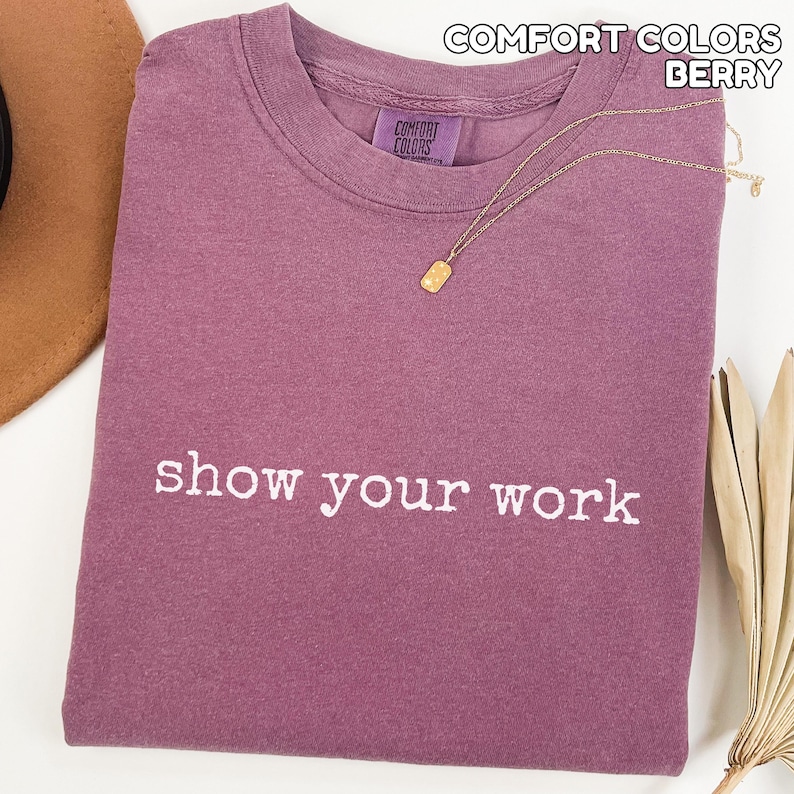 K&ouml;nnte beinhalten: Beerenfarbenes Comfort Colors T-Shirt mit dem wei&szlig;en Schriftzug "show your work". Eine goldene Halskette mit rechteckigem Anh&auml;nger liegt auf dem Shirt. Das Shirt ist ordentlich gefaltet, mit Hut und getrockneter Pflanze im Hintergrund.