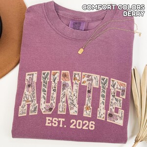 Puede incluir: Sudadera "Auntie" de color baya con letras florales. La palabra "AUNTIE" est&aacute; impresa en la parte delantera, con "EST. 2026" debajo. Un collar de oro con un colgante descansa sobre la camisa. Fabricado por Comfort Colors.