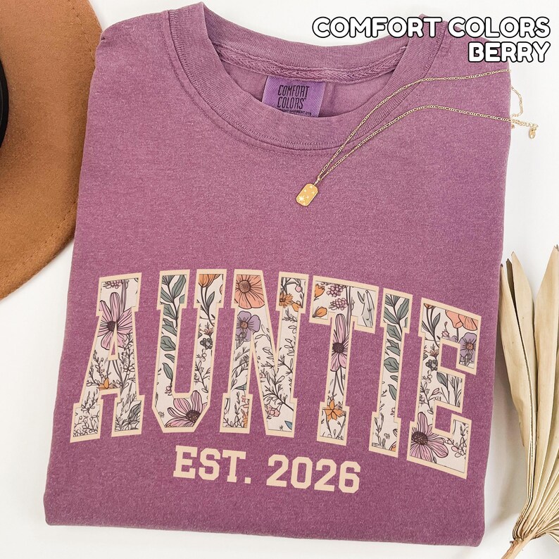 Puede incluir: Sudadera "Auntie" de color baya con letras florales. La palabra "AUNTIE" est&aacute; impresa en la parte delantera, con "EST. 2026" debajo. Un collar de oro con un colgante descansa sobre la camisa. Fabricado por Comfort Colors.