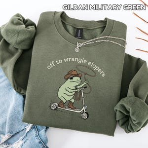 May include: Military green sweatshirt with the text "off to wrangle elopers" and a cartoon frog wearing a cowboy hat and boots, riding a scooter. A silver necklace is draped across the sweatshirt. The sweatshirt is folded next to a pair of blue jeans.