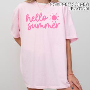 K&ouml;nnte beinhalten: Hellrosa T-Shirt mit dem Schriftzug "hello summer" in Pink und einer pinkfarbenen Sonnengrafik. Der Text "COMFORT COLORS BLOSSOM" ist oberhalb des Designs aufgedruckt. Das Shirt hat kurze &Auml;rmel.