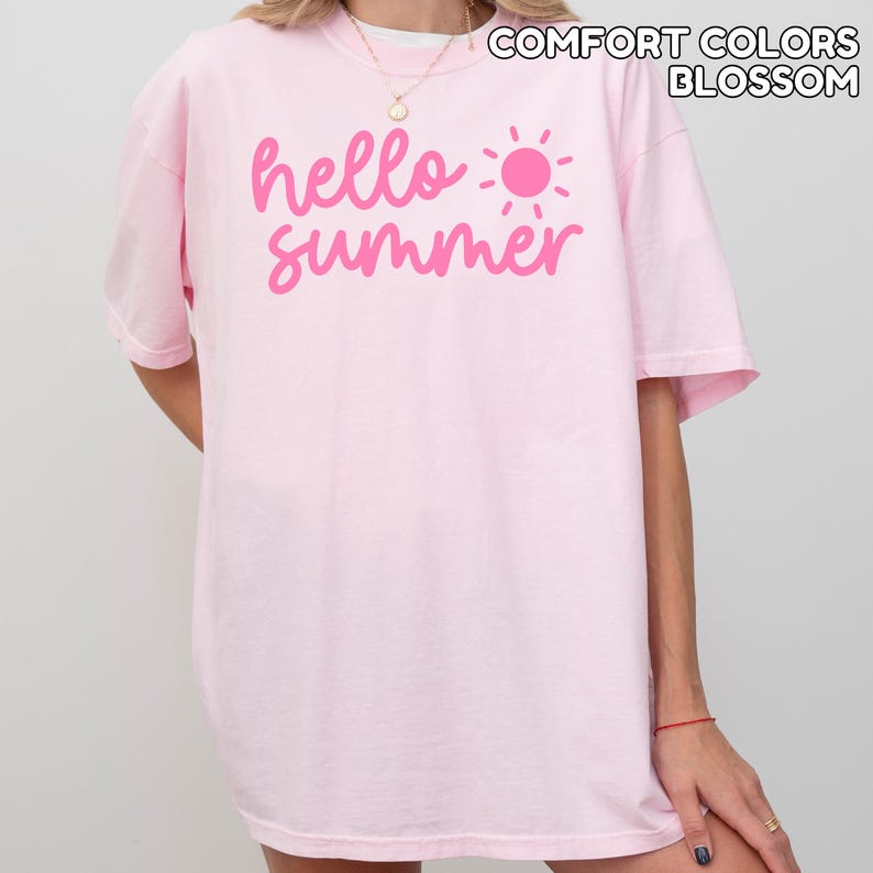 K&ouml;nnte beinhalten: Hellrosa T-Shirt mit dem Schriftzug "hello summer" in Pink und einer pinkfarbenen Sonnengrafik. Der Text "COMFORT COLORS BLOSSOM" ist oberhalb des Designs aufgedruckt. Das Shirt hat kurze &Auml;rmel.