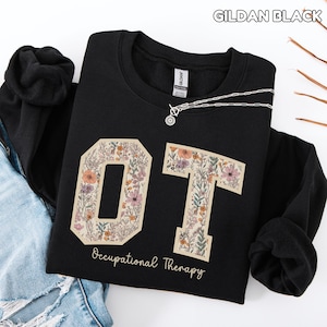 Może przedstawiać: Czarna bluza z literami "OT" w kwiatowym wzorze. Poniżej napis "Occupational Therapy". Srebrny naszyjnik z wisiorkiem jest zarzucony na bluzę.