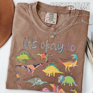 May include: Espresso-colored t-shirt with the phrase "it's okay to" and colorful dinosaur illustrations. The shirt has a crew neck and short sleeves. A silver necklace with a pendant is draped on the shirt.