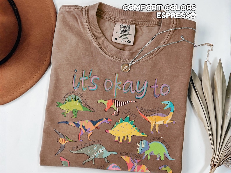 May include: Espresso-colored t-shirt with the phrase "it's okay to" and colorful dinosaur illustrations. The shirt has a crew neck and short sleeves. A silver necklace with a pendant is draped on the shirt.