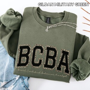 May include: A military green Gildan crewneck sweatshirt featuring the black text BCBA with a leopard print outline. Below the letters, the text Board Certified Behavior Analyst is printed in a cursive font. A silver chain necklace rests on the collar.