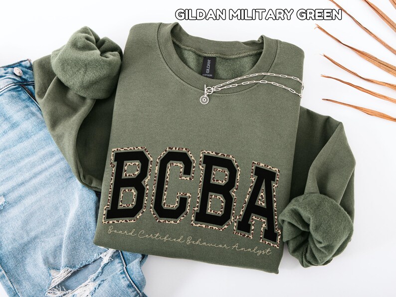 May include: A military green Gildan crewneck sweatshirt featuring the black text BCBA with a leopard print outline. Below the letters, the text Board Certified Behavior Analyst is printed in a cursive font. A silver chain necklace rests on the collar.