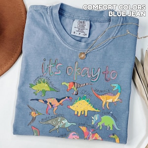 May include: Blue jean Comfort Colors t-shirt with a crew neck and short sleeves. The shirt features colorful dinosaur illustrations and the phrase "it's okay to" with other words. A gold necklace is draped across the shirt.
