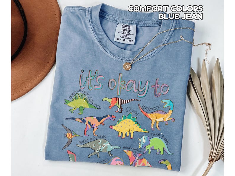 May include: Blue jean Comfort Colors t-shirt with a crew neck and short sleeves. The shirt features colorful dinosaur illustrations and the phrase "it's okay to" with other words. A gold necklace is draped across the shirt.