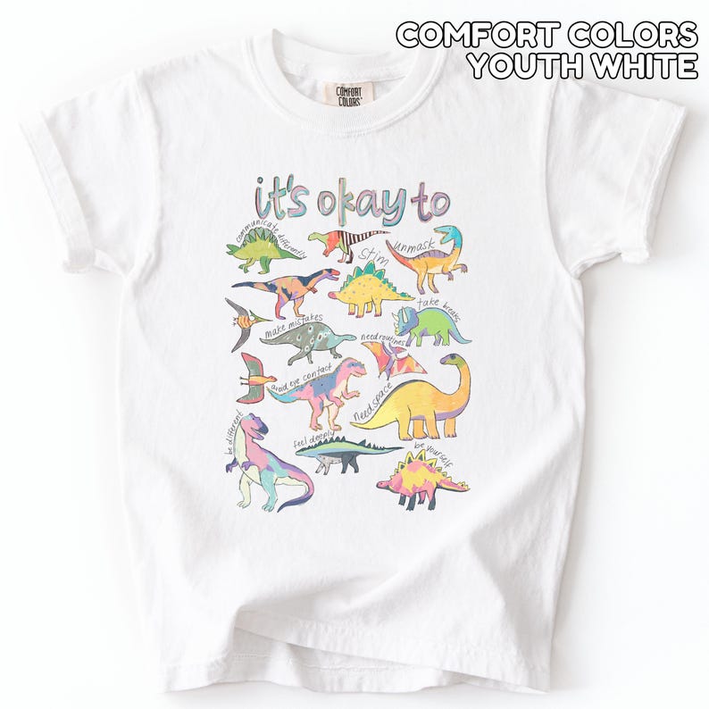 May include: White youth t-shirt with a colorful dinosaur graphic. The shirt features the phrase "it's okay to" above various dinosaur illustrations. The dinosaurs are in various colors and poses, with encouraging words.