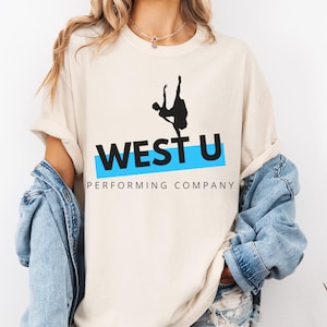 May include: Cream-colored t-shirt featuring the text "WEST U" in black letters with a light blue horizontal stripe. A black silhouette of a dancer is above the text. The words "PERFORMING COMPANY" are below the text.
