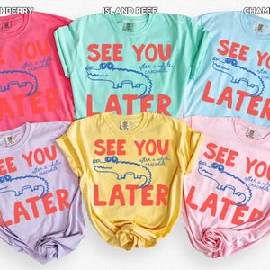 May include: A selection of t-shirts in various colours, including pink, blue, green, yellow, and purple. Each shirt displays the phrase "SEE YOU LATER" alongside a cartoon crocodile illustration. The text and crocodile are in contrasting colours.