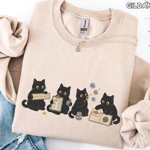 May include: A sand-colored crewneck sweatshirt featuring a graphic of four black cats. One cat holds a star-rated checklist, another a clipboard, the third blows bubbles, and the fourth examines a book with a fish and yarn illustration.