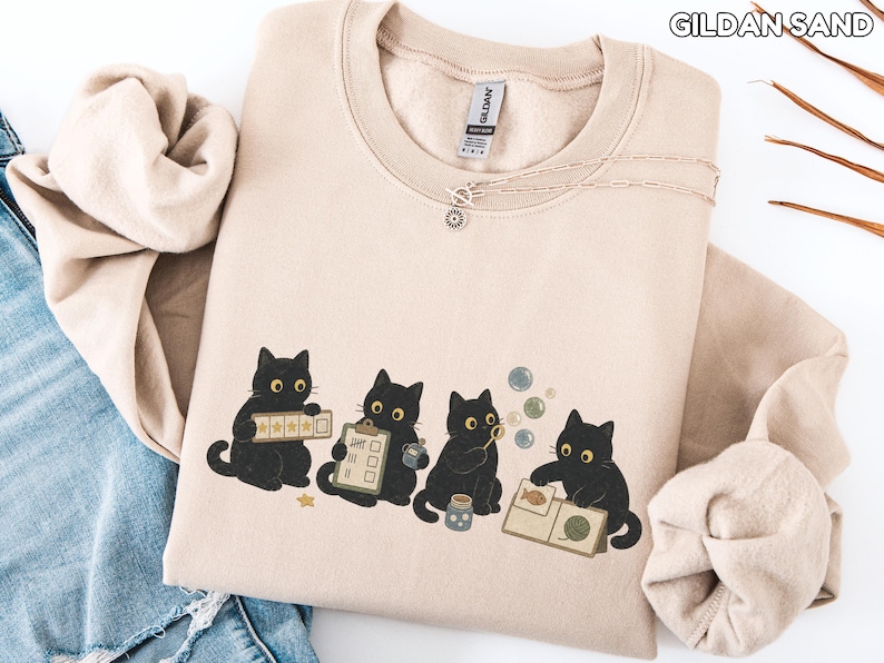May include: A sand-colored crewneck sweatshirt featuring a graphic of four black cats. One cat holds a star-rated checklist, another a clipboard, the third blows bubbles, and the fourth examines a book with a fish and yarn illustration.