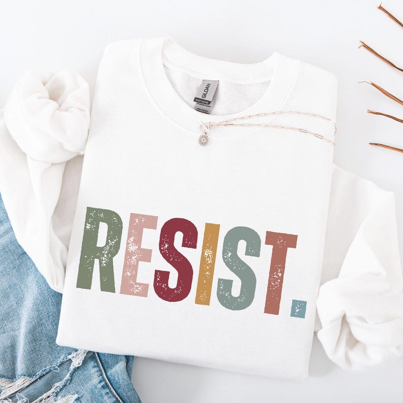 Pode incluir: Camisola branca com a palavra "RESIST" impressa em letras coloridas e desgastadas. As letras s&atilde;o em tons de verde, rosa, vermelho, amarelo e azul.