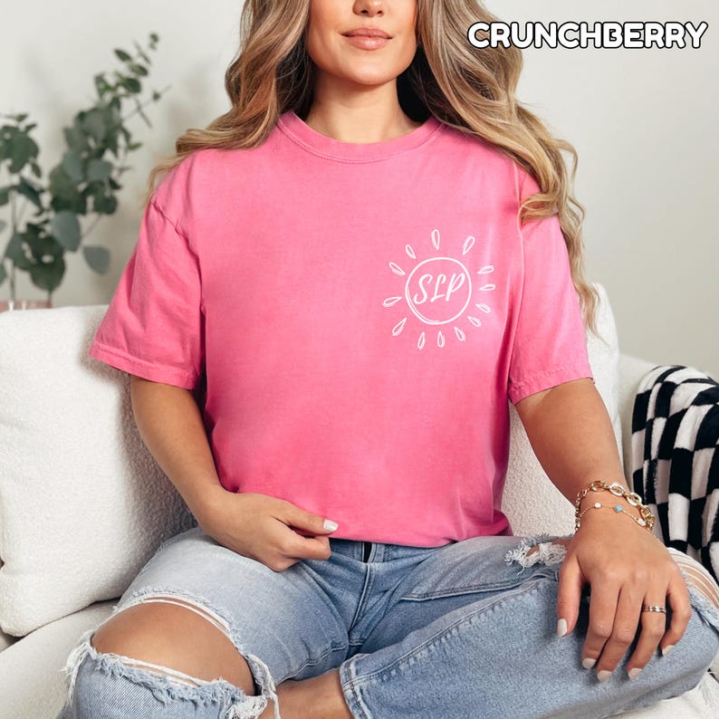 May include: A pink t-shirt with the word "CRUNCHBERRY" at the top. The shirt has a white sun design with the letters "SLP" inside. The person is wearing blue jeans.