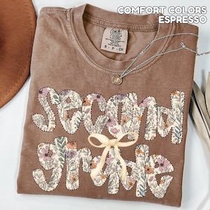 May include: Espresso-colored t-shirt with the words "Second Grade" in a floral design. The shirt has a crew neck and short sleeves. A silver necklace with a pendant and a cream-colored bow are on the shirt.