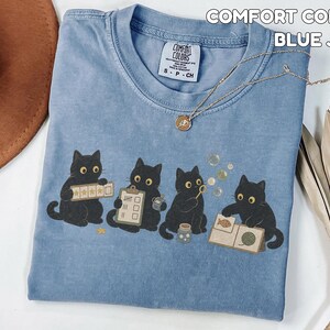 May include: A blue jean colored Comfort Colors t-shirt featuring a graphic of four black cats. The cats are depicted interacting with various items including a star rating board, a clipboard, a bubble wand, and a book with a yarn ball.