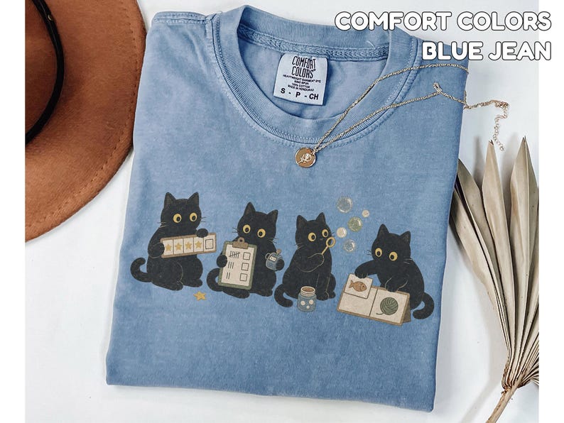 May include: A blue jean colored Comfort Colors t-shirt featuring a graphic of four black cats. The cats are depicted interacting with various items including a star rating board, a clipboard, a bubble wand, and a book with a yarn ball.