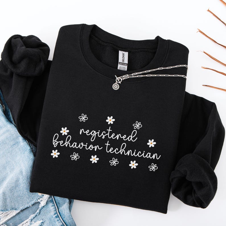 K&ouml;nnte beinhalten: Schwarzer Pullover mit dem Text "registered behavior technician" in wei&szlig;er Schrift mit kleinen wei&szlig;en G&auml;nsebl&uuml;mchen um den Text herum.