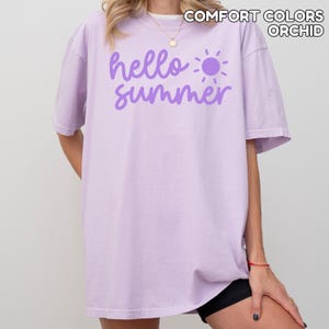 K&ouml;nnte beinhalten: Ein helllila Comfort Colors T-Shirt mit dem Schriftzug "hello summer" in lila Schrift und einer lila Sonnengrafik. Das Shirt hat eine lockere Passform und kurze &Auml;rmel.