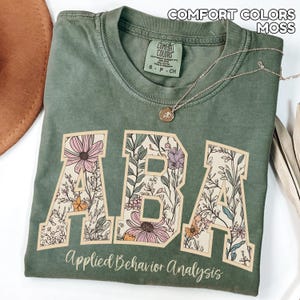 May include: A moss green t-shirt with the letters "ABA" in a floral design. The words "Applied Behavior Analysis" are printed below the letters. A gold necklace with a pendant is draped across the shirt.