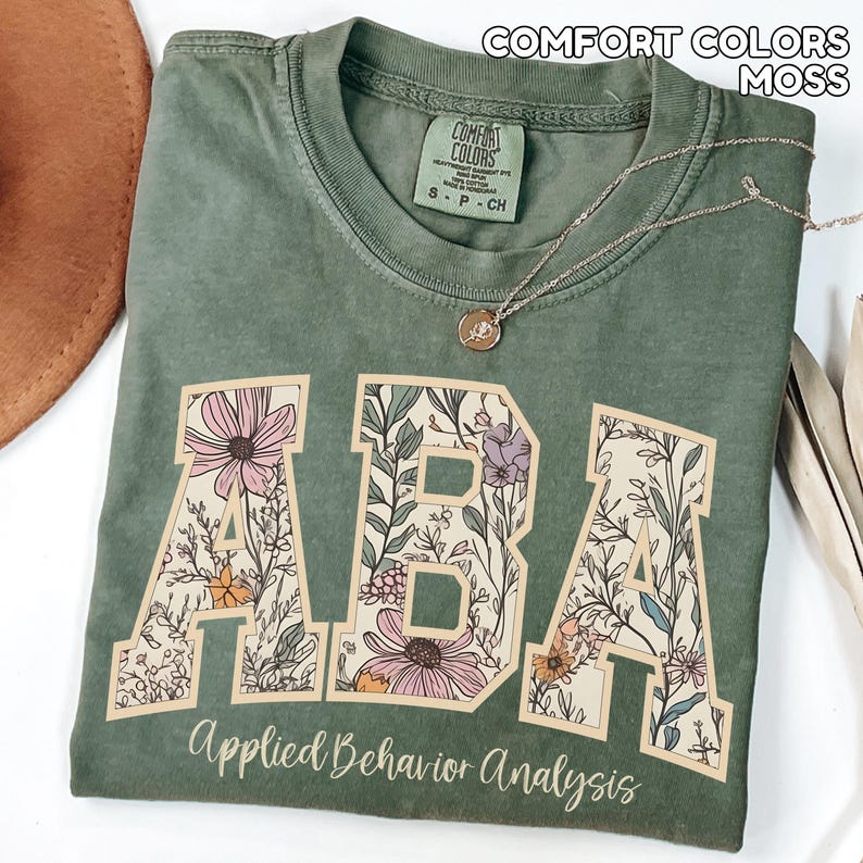 May include: A moss green t-shirt with the letters "ABA" in a floral design. The words "Applied Behavior Analysis" are printed below the letters. A gold necklace with a pendant is draped across the shirt.