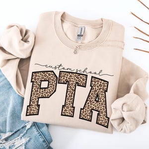 May include: A beige sweatshirt with the text "custom school PTA" in black script and leopard print block letters. A silver necklace with a small pendant is draped on the sweatshirt. The sweatshirt is folded on a white surface.
