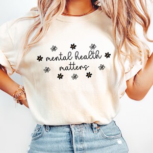May include: A white t-shirt with the text "mental health matters" in black script with small black flowers around the text.