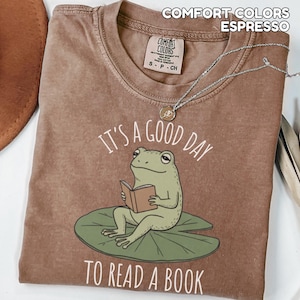 May include: Espresso-colored t-shirt with the text "IT'S A GOOD DAY TO READ A BOOK" and a cartoon frog reading a book while sitting on a lily pad. A silver necklace with a pendant is on the shirt.